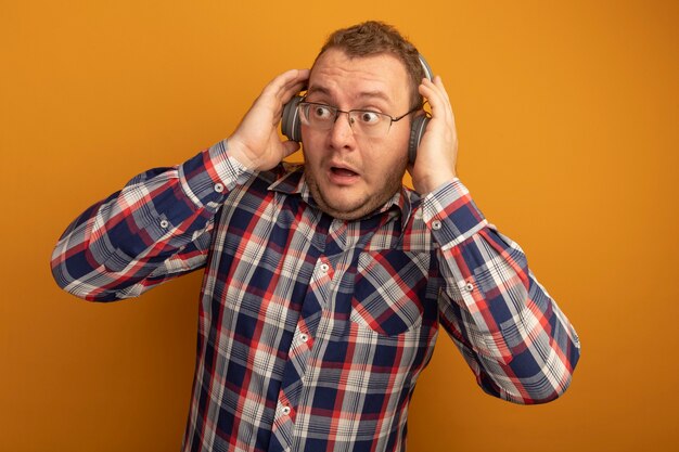 Hombre de gafas y camisa a cuadros con auriculares mirando a un lado confundido parado sobre pared naranja