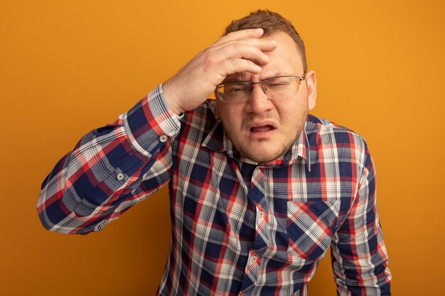 Hombre de gafas y camisa a cuadros confundido con la mano sobre la cabeza frunciendo el ceño de pie sobre la pared naranja