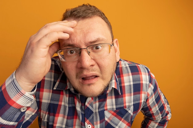 Hombre de gafas y camisa a cuadros confundido y preocupado con la mano en la cabeza de pie sobre la pared naranja