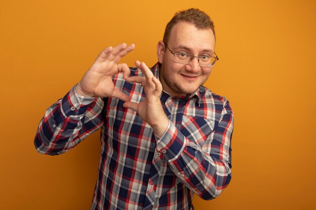 Hombre con gafas y camisa a cuadros haciendo gesto de corazón con los dedos sonriendo alegremente de pie sobre la pared naranja