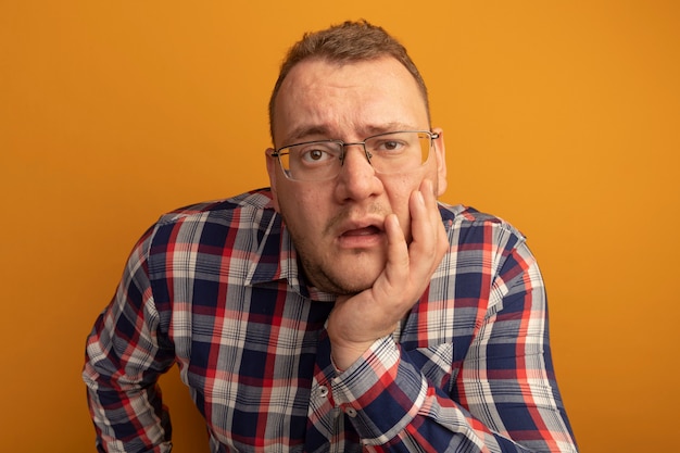 Hombre de gafas y camisa a cuadros con la mano en el rostro confundido de pie sobre la pared naranja