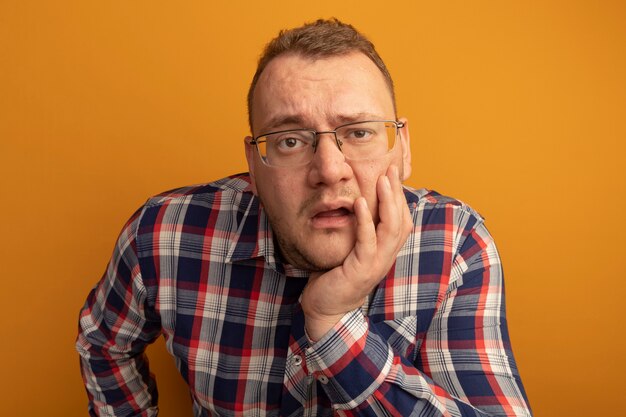 Hombre de gafas y camisa a cuadros con la mano en el rostro confundido de pie sobre la pared naranja