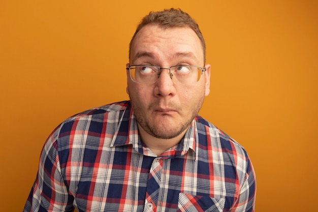 Hombre de gafas y camisa a cuadros mirando hacia arriba preocupado y confundido de pie sobre la pared naranja