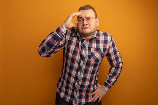 Hombre de gafas y camisa a cuadros mirando a un lado confundido con la mano en la cabeza de pie sobre la pared naranja