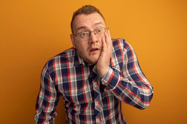 Hombre con gafas y camisa a cuadros mirando a un lado confundido y muy ansioso con la mano en la cara de pie sobre la pared naranja