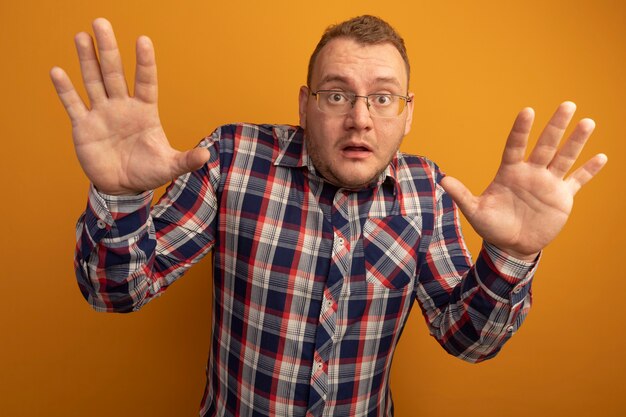 Hombre de gafas y camisa a cuadros preocupado con las manos levantadas en rendición de pie sobre la pared naranja