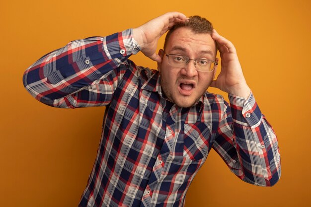 Hombre de gafas y camisa a cuadros siendo señalado con las manos en la cabeza de pie sobre la pared naranja