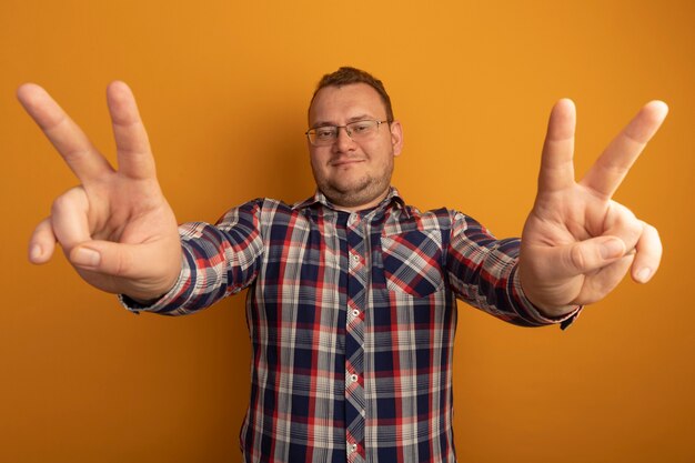 Hombre de gafas y camisa a cuadros sonriendo alegremente mostrando v-sign de pie sobre la pared naranja