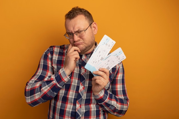 Hombre con gafas y camisa a cuadros sosteniendo boletos de avión mirando a un lado con la mano en la barbilla, cejas fruncidas, disgustado de pie sobre la pared naranja