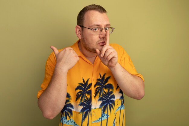 Hombre de gafas con camisa naranja haciendo gesto de silencio con el dedo en los labios de pie sobre la pared de luz