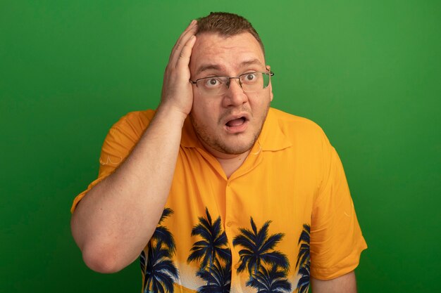 Hombre de gafas y camisa naranja mirando a un lado confundido con la mano en la cabeza por error de pie sobre la pared verde