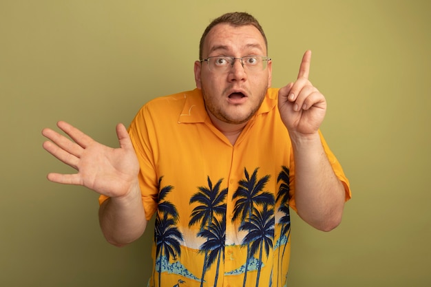 Hombre de gafas con camisa naranja mirando sorprendido mostrando el dedo índice de pie sobre la pared de luz