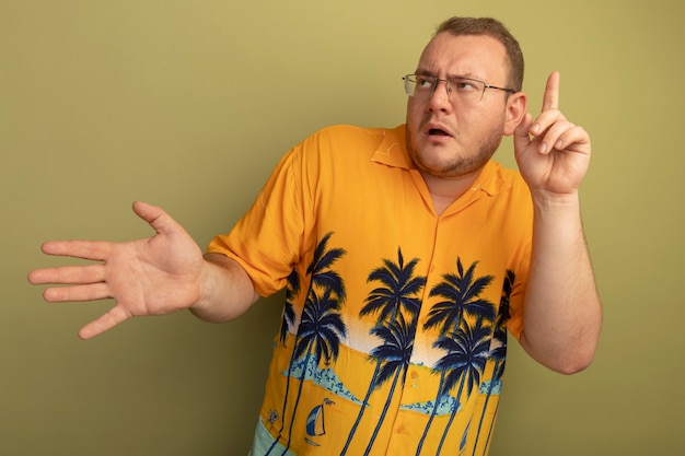 Hombre de gafas con camisa naranja que parece confundido mostrando la palma abierta y el dedo índice de pie sobre una pared de luz