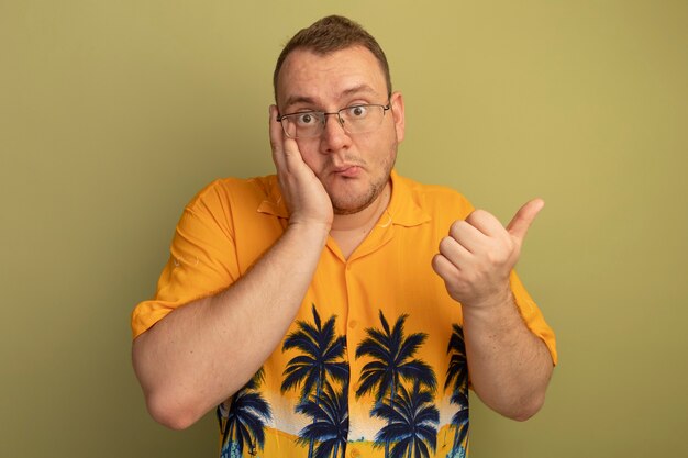 Hombre de gafas con camisa naranja que parece confundido mostrando los pulgares para arriba confundido de pie sobre la pared de luz