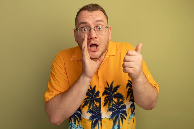 Hombre de gafas con camisa naranja sorprendido apuntando hacia atrás sosteniendo la mano cerca de la boca de pie sobre una pared ligera