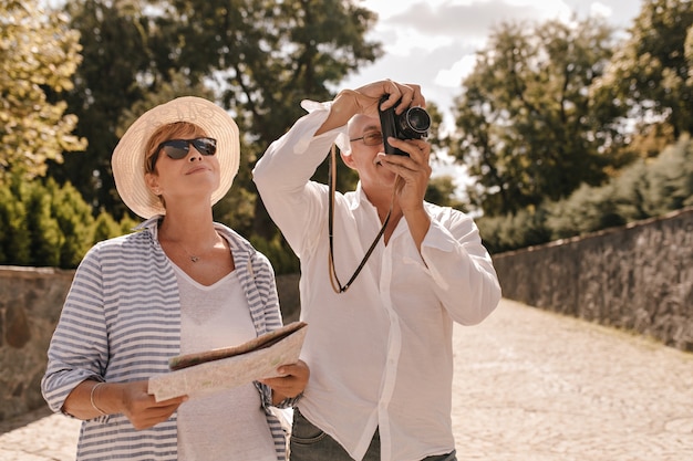 Hombre con gafas y fotografías de camisa blanca elegante y sonríe con una dama rubia con gafas de sol, sombrero y ropa fresca a rayas con mapa al aire libre.