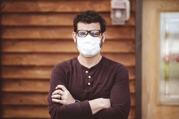 Hombre con gafas y una mascarilla sanitaria con los brazos cruzados delante de una pared de madera