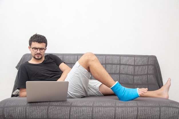 Hombre con gafas con pierna rota en férula azul para el tratamiento de lesiones por esguince de tobillo trabajando en una computadora portátil en el sofá en casa.