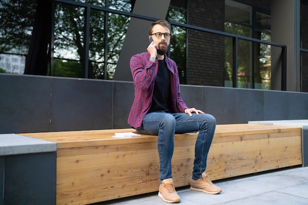 Hombre con gafas sentado en un banco de madera y hablando por teléfono