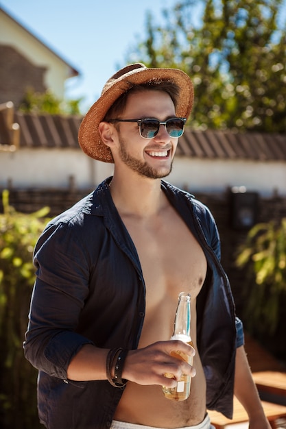 Hombre con gafas de sol y sombrero bebiendo cerveza, descansando en casa de campo