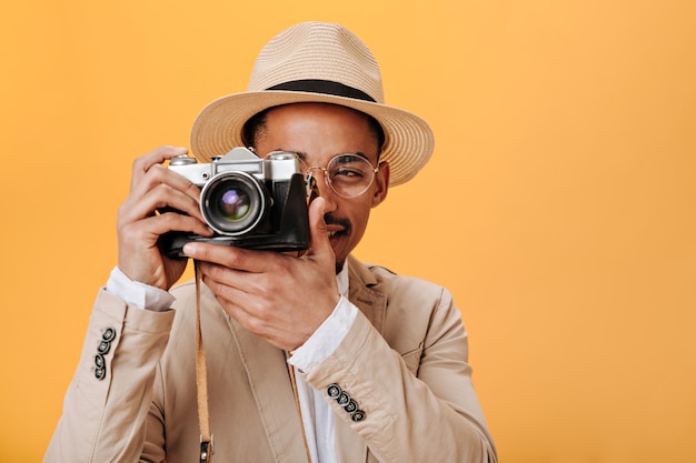 Hombre con gafas y sombrero posando en la pared naranja con cámara retro
