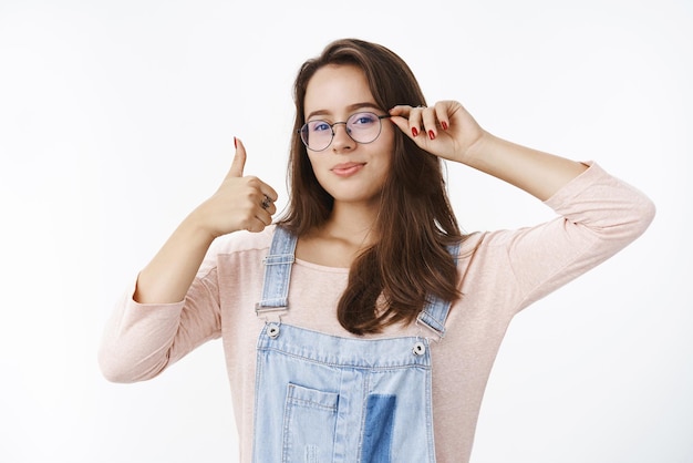 hombre con gafas y todos los labios doblados, lindo y sonriente, mostrando los pulgares hacia arriba en señal de aprobación y de acuerdo, gustando la idea y dando recomendaciones sobre la pared gris.