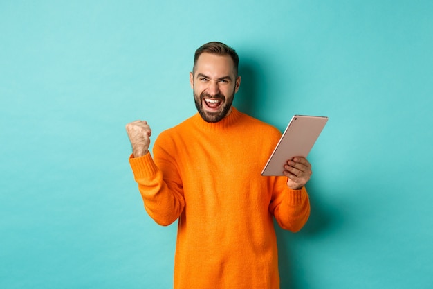 Hombre ganador alegre que sostiene la tableta digital, regocijándose y celebrando la victoria en el juego, haciendo el puño