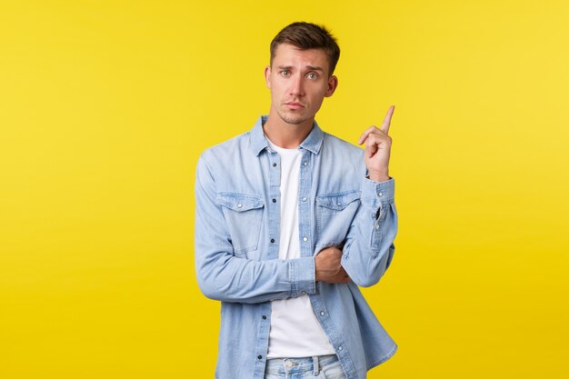 Hombre gay guapo de aspecto serio con camisa de mezclilla, levantando el dedo índice tiene una sugerencia, diciendo su idea o plan, encontró una buena solución, de pie con un fondo amarillo determinado.