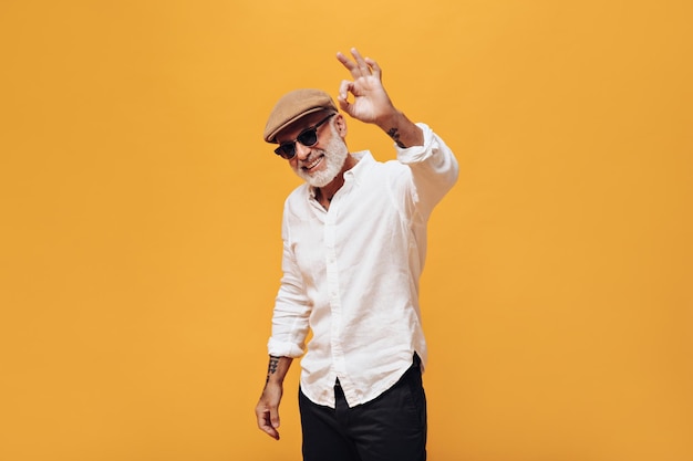 Un hombre genial con un elegante sombrero de camisa y gafas de sol que muestran el signo ok Un tipo de pelo gris con gorra beige y una camisa ligera sonríe y se divierte