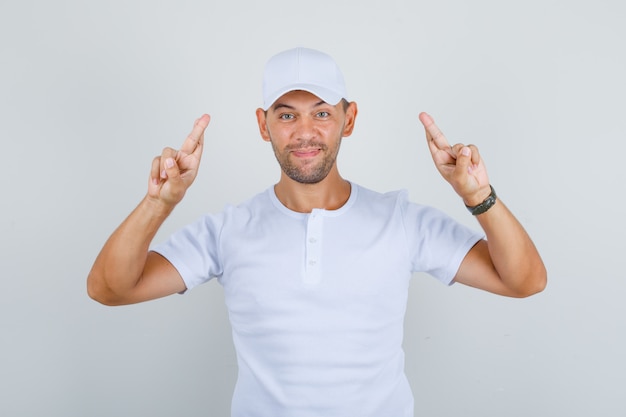 Hombre gesticulando con el dedo cruzado y deseando suerte en camiseta blanca, gorra y mirando feliz, vista frontal.