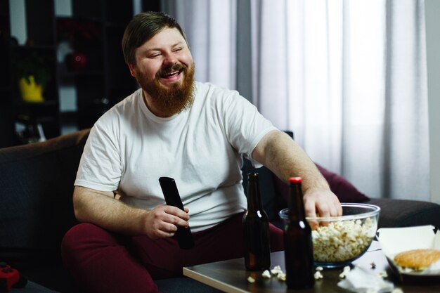 El hombre gordo feliz se sienta en el sofá y mira la TV con palomitas de maíz y cerveza