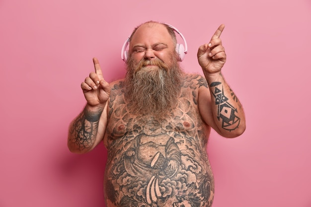 Hombre gordo relajado y divertido con cuerpo desnudo, brazos y estómago tatuados, baila mientras escucha música, mueve los brazos y cierra los ojos con alegría, usa audífonos en las orejas, se divierte y siente aspiración