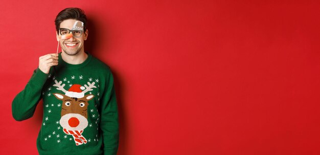 Hombre gracioso con suéter de Navidad y máscara de fiesta, celebrando las vacaciones de invierno, sonriendo feliz, de pie sobre un fondo rojo.