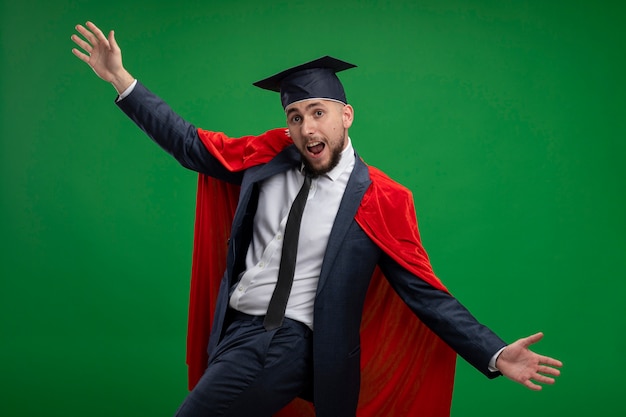 Hombre graduado en capa roja feliz y emocionada amplia apertura manos de pie sobre la pared verde