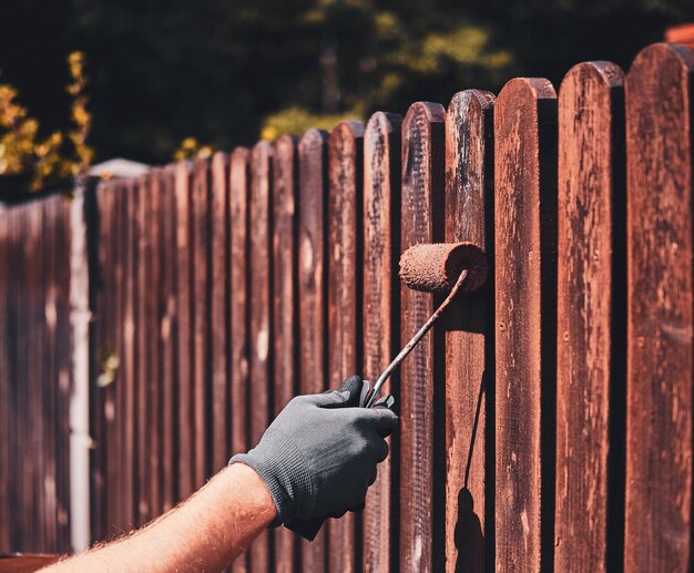 El hombre con guantes protectores está pintando una cerca de madera en un brillante día de verano.