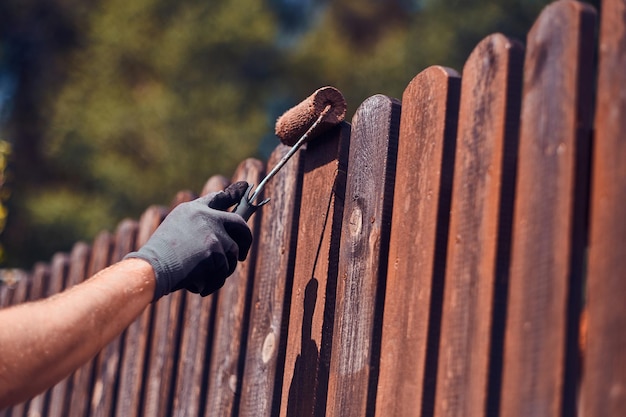 El hombre con guantes protectores está pintando una cerca de madera en un brillante día de verano.