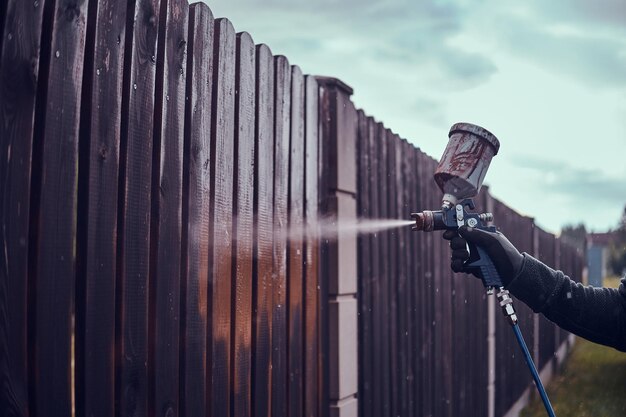 El hombre y los guantes protectores están aplicando pintura a la antigua valla usando aerógrafo en su jardín.