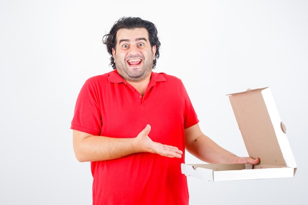 Hombre guapo abriendo la caja de papel, estirando la mano hacia él con una manera feliz en camiseta roja y luciendo alegre. vista frontal.