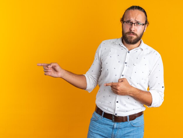 Hombre guapo adulto con el ceño fruncido con gafas apuntando al lado