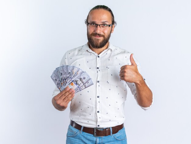 Hombre guapo adulto complacido con gafas mostrando dinero y pulgar hacia arriba mirando a cámara aislada en la pared blanca