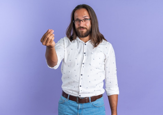 Hombre guapo adulto confiado con gafas mirando a la cámara haciendo gesto de dinero aislado en la pared púrpura