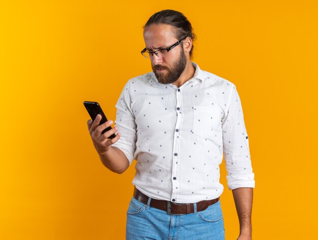 Hombre guapo adulto confundido con gafas sosteniendo y mirando el teléfono móvil aislado en la pared naranja