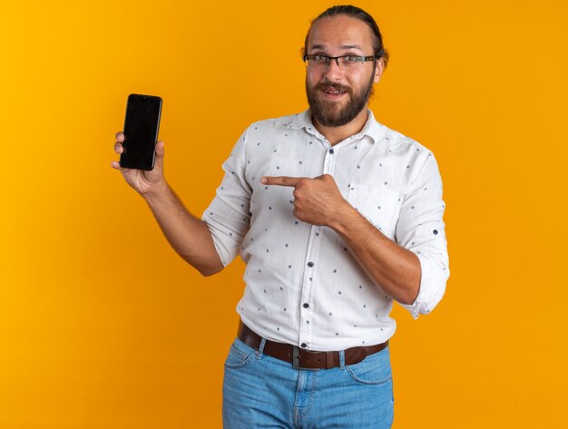 Hombre guapo adulto emocionado con gafas mostrando teléfono móvil apuntando a él mirando a la cámara aislada en la pared naranja