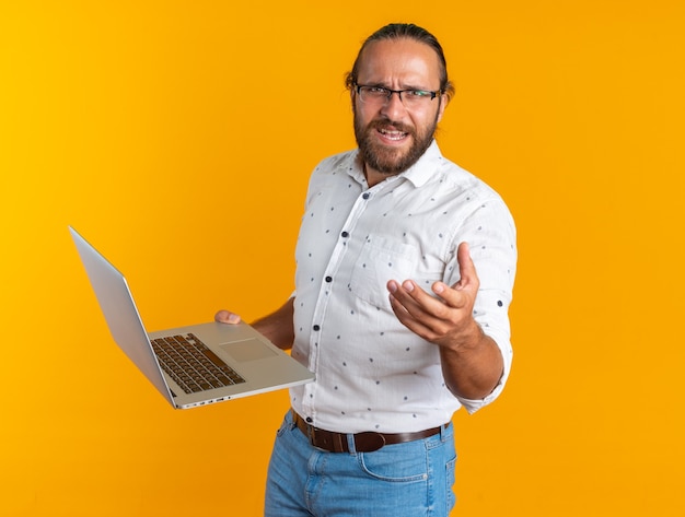 Hombre guapo adulto enojado con gafas de pie en la vista de perfil sosteniendo portátil mostrando la mano vacía