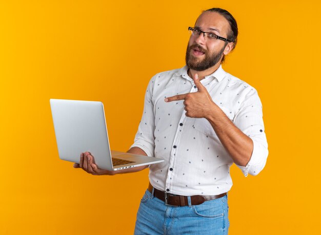 Hombre guapo adulto impresionado con gafas sosteniendo y apuntando a la computadora portátil