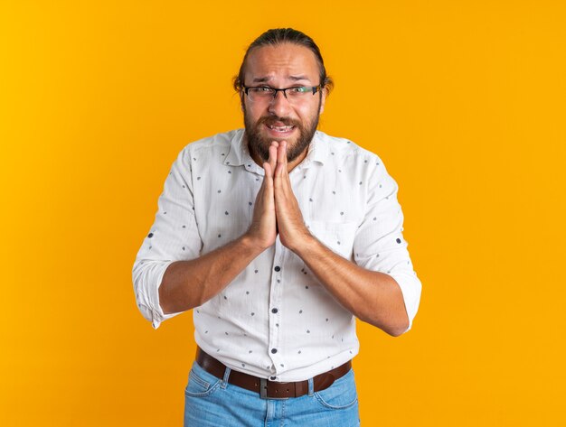 Hombre guapo adulto nervioso con gafas mirando a la cámara manteniendo las manos juntas orando aislado en la pared naranja