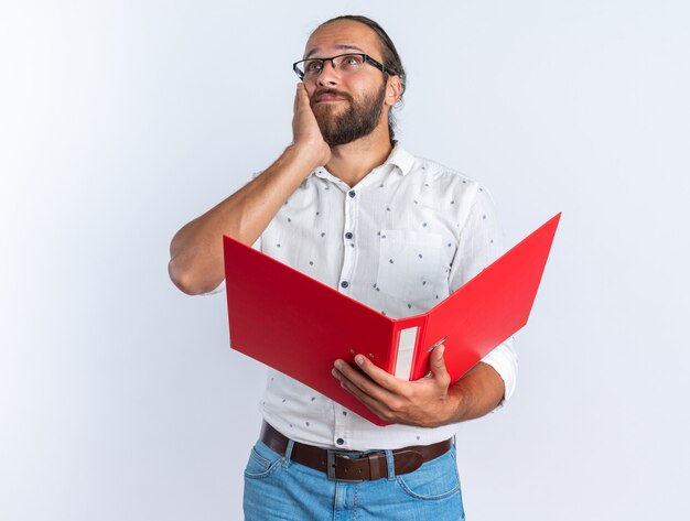 Hombre guapo adulto pensativo con gafas manteniendo la mano en la cara sosteniendo la carpeta abierta mirando al lado aislado en la pared blanca