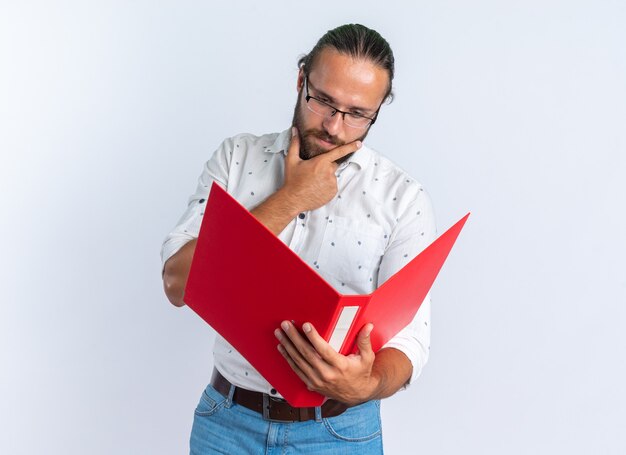 Hombre guapo adulto pensativo con gafas sosteniendo y mirando la carpeta manteniendo la mano en la barbilla aislada en la pared blanca