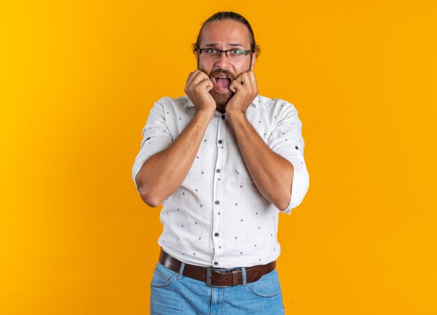Hombre guapo adulto preocupado con gafas manteniendo las manos en la cara mirando a la cámara con la boca abierta aislada en la pared naranja con espacio de copia