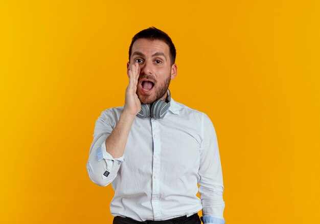 Hombre guapo alegre con auriculares en el cuello sostiene la mano cerca de la boca pretendiendo llamar a alguien aislado en la pared naranja